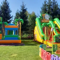 Dwa kolorowe, dmuchane zamki w stylu dżungli z motywami zwierzęcymi, ustawione na trawniku w słoneczny dzień, otoczone kolorowym płotkiem z tworzywa sztucznego.