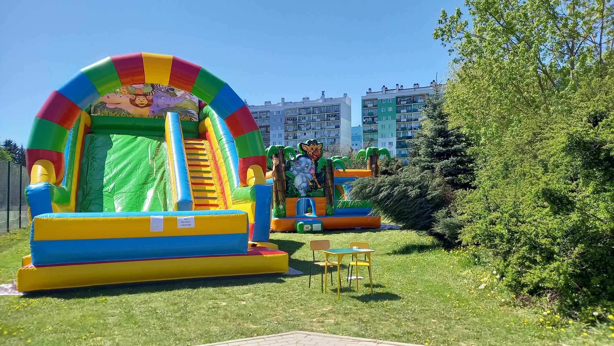 Dwa kolorowe, dmuchane place zabaw z motywami zwierząt, ustawione na trawniku przed budynkami mieszkalnymi, obok stolik z krzesłami.