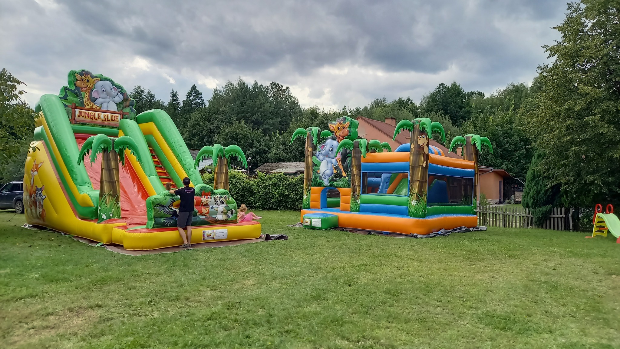 Dwa kolorowe dmuchane zamki w dżungli na trawie, z motywami zwierzęcymi i palmami, jeden ze zjeżdżalnią, a drugi z małym basenem. Osoba nadzoruje zamek ze zjeżdżalnią.