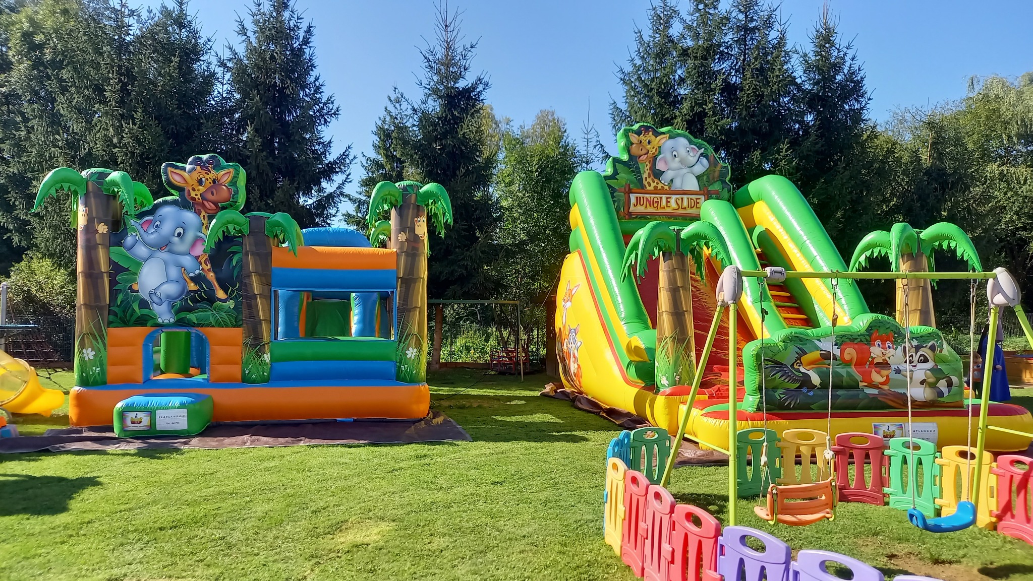 Dwa kolorowe, dmuchane zamki w stylu dżungli z motywami zwierzęcymi, ustawione na trawniku w słoneczny dzień, otoczone kolorowym płotkiem z tworzywa sztucznego.