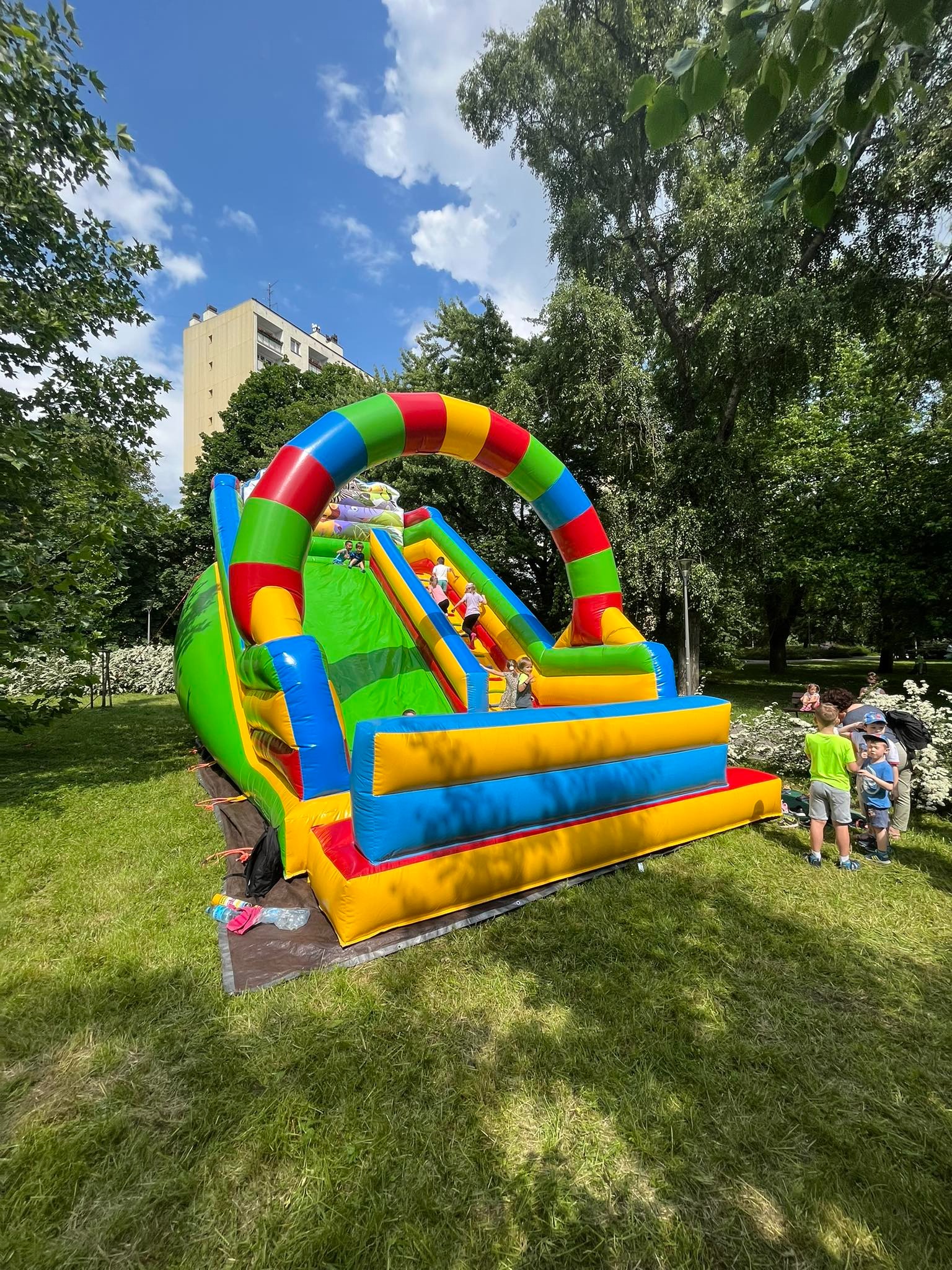 Kolorowy dmuchany zamek ze zjeżdżalnią w parku, dzieci zjeżdżają w dół, a w tle widać budynek i drzewa.
