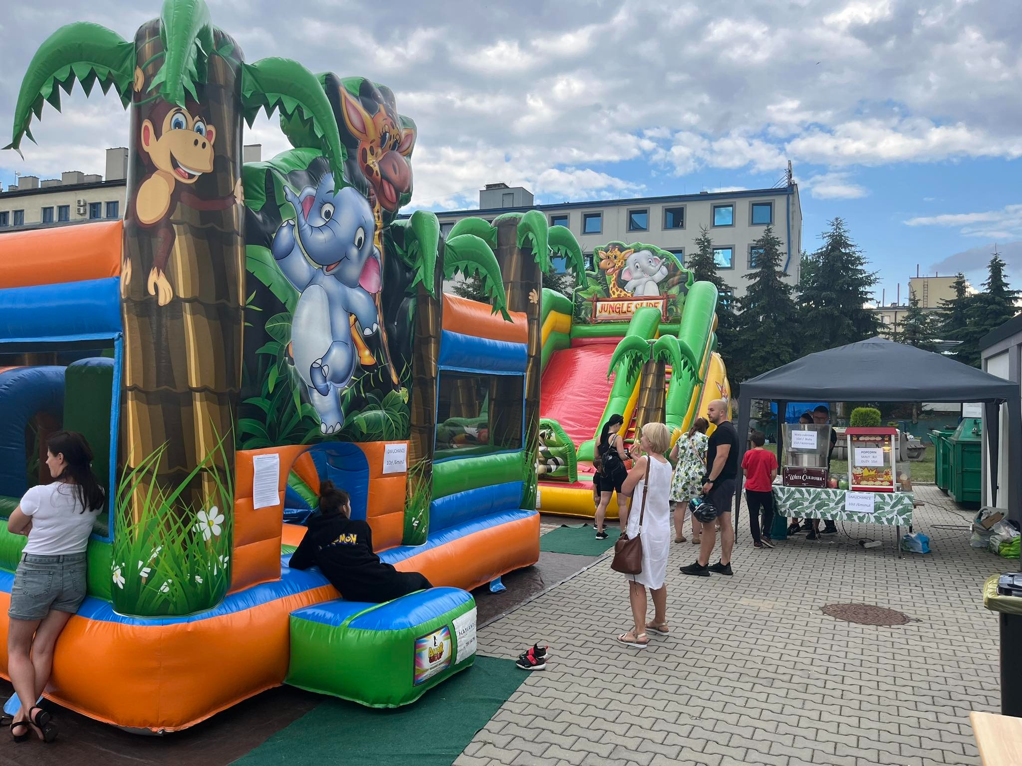 Dwa dmuchane zamki w stylu dżungli z motywami zwierzęcymi, ustawione na zewnątrz podczas imprezy plenerowej, z osobami w tle i stoiskiem z watą cukrową widocznym po prawej stronie.