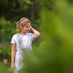 Fotorama24 - Dziewczynka w białej sukience komunijnej z perłową opaską na głowie, pozująca na tle zieleni.