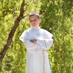 Fotorama24 - Chłopiec w białej albie ze złotymi zdobieniami, z założonymi rękami, pozuje na tle zieleni drzew. Uśmiech na twarzy, naturalne światło.