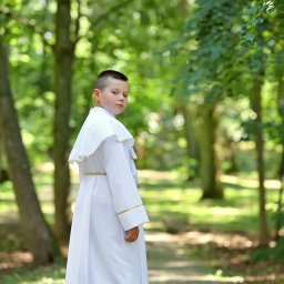 Fotorama24 - Chłopiec w białej albie komunijnej z złotym sznurkiem, spogląda przez ramię w obiektyw na tle zielonego parku w Ciechanowie.