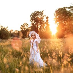 Fotorama24 - Dziewczynka w białej sukience i z koronkowym parasolem stoi na łące w promieniach zachodzącego słońca. Włosy długie, proste, wianek na głowie. Delikatne, rozproszone światło podkreśla naturalny...