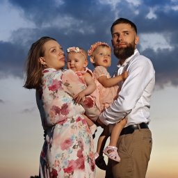 Fotorama24 - Rodzina z dwiema małymi córeczkami w pastelowych sukienkach i kwiatowych opaskach, pozująca na tle zachmurzonego nieba o zmierzchu.