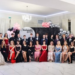 Fotorama24 - Grupowe zdjęcie uczniów i uczennic na tle dekoracji studniówkowej, z napisem 'Studniówka 2024', balonami i błyszczącą ścianką, w eleganckim wnętrzu z marmurową podłogą i kryształowym żyrandolem.