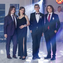 Fotorama24 - Grupa młodych, elegancko ubranych osób pozuje na tle sceny z oświetleniem eventowym, w tym reflektorami i podświetlanym napisem.