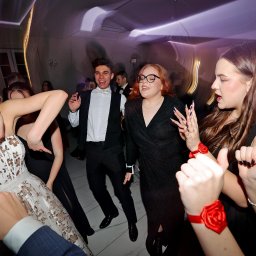 Fotorama24 - Rozbawieni goście w tańcu podczas balu, widoczna sukienka z haftem, garnitur z muszką i czerwone ozdoby na nadgarstkach.