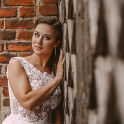 Fotorama24 - Portret kobiety w koronkowej sukni, opierającej się o ścianę z cegły i kamienia, z delikatnym makijażem i długimi kolczykami.