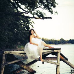 Fotorama24 - Uśmiechnięta kobieta w białej sukni i z wiankiem na głowie siedzi na drewnianym pomoście nad jeziorem, z drzewami w tle.