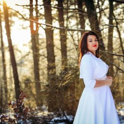 Fotorama24 - Kobieta w białej sukni i bolerku pozuje w zimowym lesie, oświetlona promieniami słońca przebijającymi przez drzewa. Czerwone usta i ciemne włosy kontrastują z bielą stroju i otoczenia.