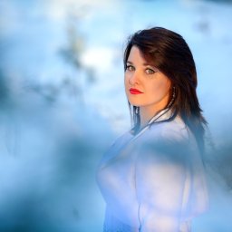 Fotorama24 - Portret kobiety z ciemnymi włosami i czerwonymi ustami, spoglądającej przez delikatną zasłonę z rozmytych gałązek na tle jasnego, błękitnego nieba.