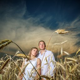 Fotorama24 - Para w białych ubraniach stoi pośród pola zboża, spoglądając w górę na dramatyczne, zachmurzone niebo. Kłosy zboża na pierwszym planie rozmyte.