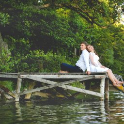 Fotorama24 - Para młoda siedząca na drewnianym pomoście nad jeziorem, opierająca się plecami o siebie, w tle bujna zieleń drzew.