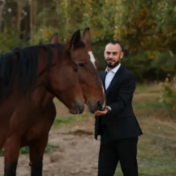 Elegancki mężczyzna w garniturze karmiący z ręki dwa brązowe konie na tle leśnej scenerii.
