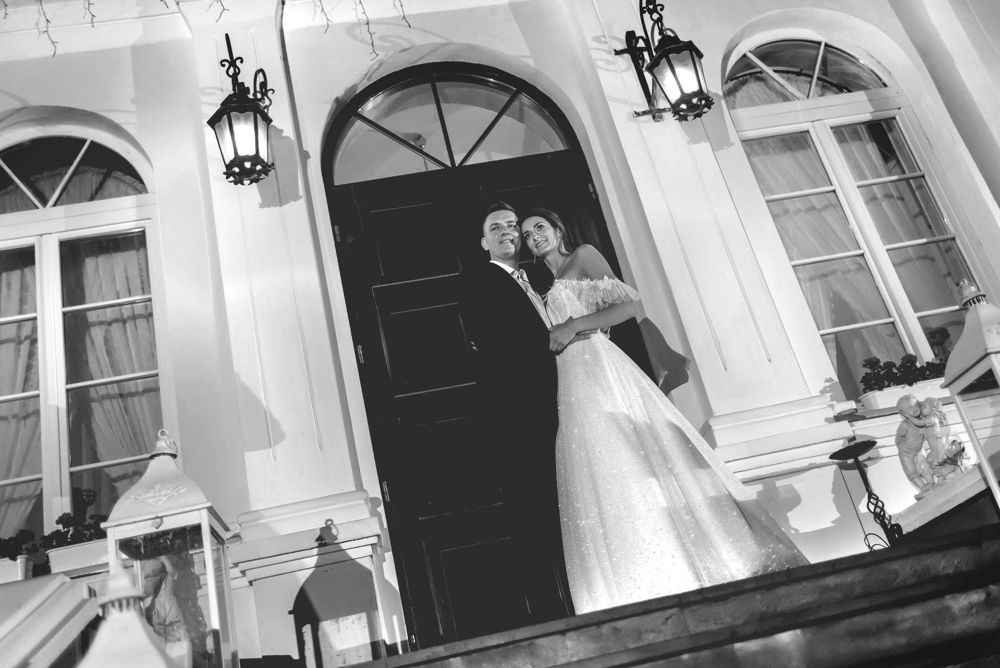 Para młoda w czerni i bieli, pozująca przed ciemnymi drzwiami z łukowym oknem, oświetlona przez ozdobne latarnie, z widocznymi detalami architektonicznymi i rzeźbami.