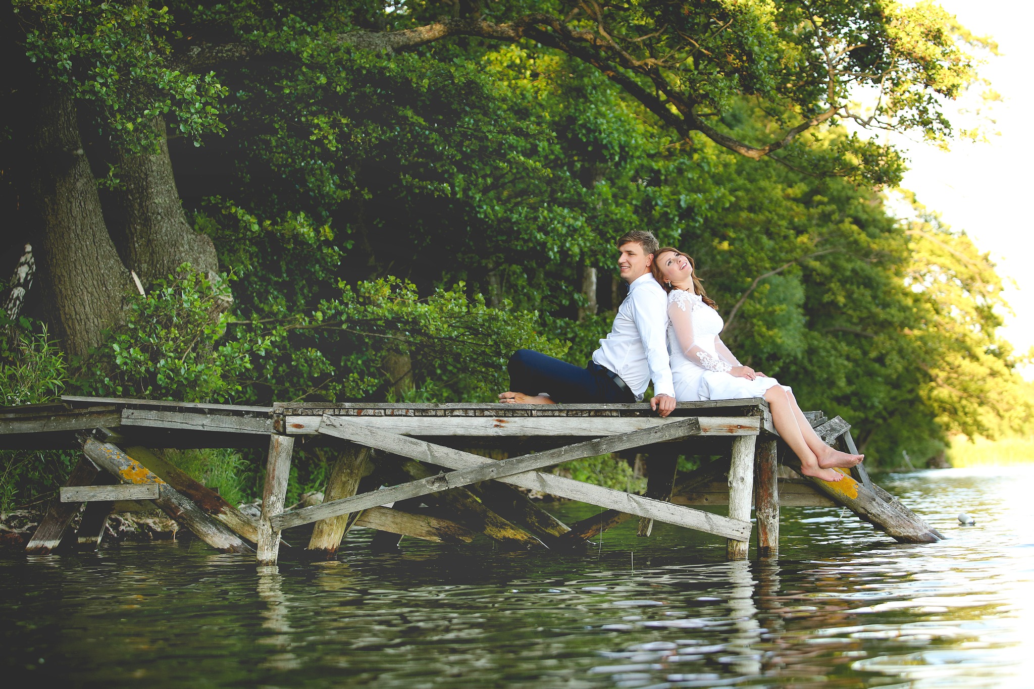 Para młoda siedząca na drewnianym pomoście nad jeziorem, opierająca się plecami o siebie, w tle bujna zieleń drzew.