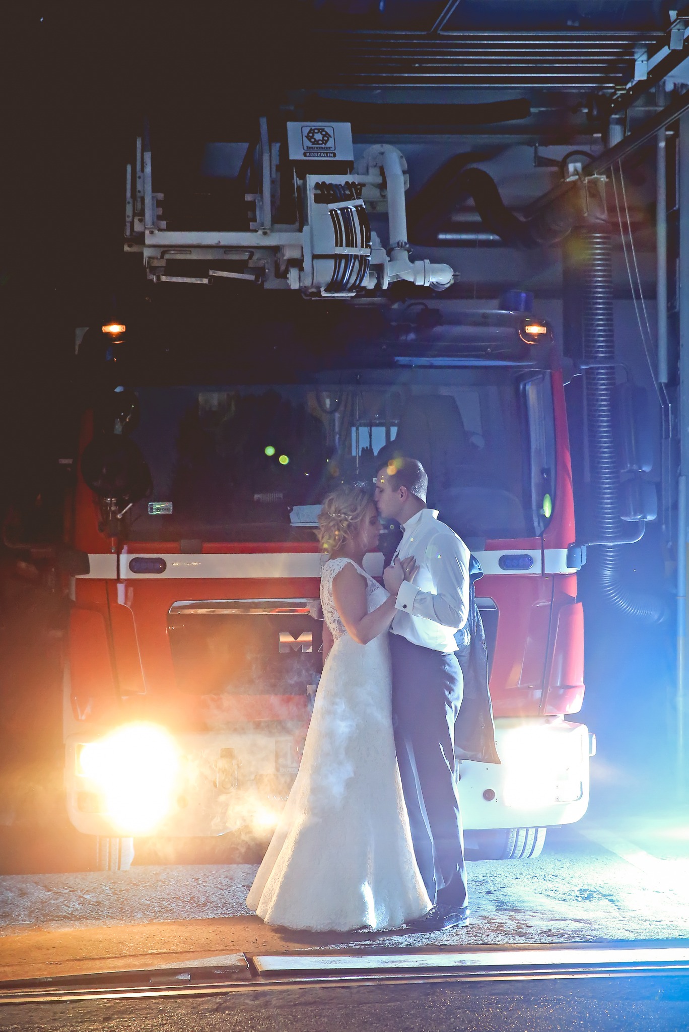 Para młoda w objęciach na tle czerwonego wozu strażackiego z włączonymi światłami, nietypowa sesja ślubna w remizie strażackiej.