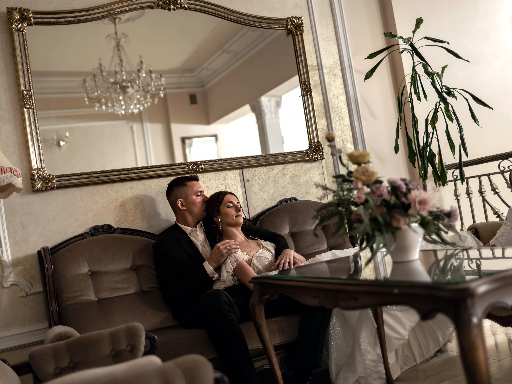 Para młoda w eleganckich strojach siedzi na sofie w bogato zdobionym wnętrzu z lustrem i żyrandolem w tle, mężczyzna całuje kobietę w czoło.