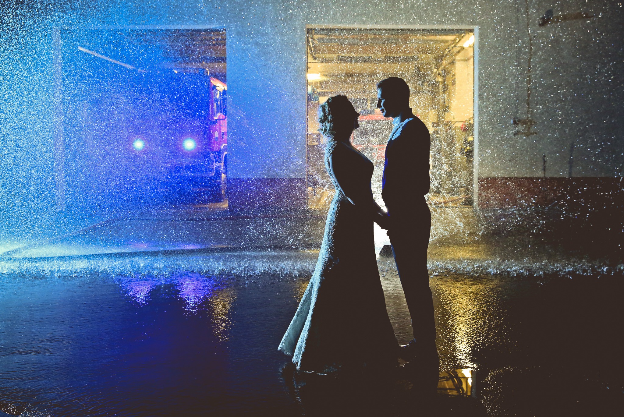 Sylwetki panny młodej w długiej sukni i pana młodego stojące pod strumieniem wody, w tle garaż strażacki z wozem strażackim oświetlonym na niebiesko.