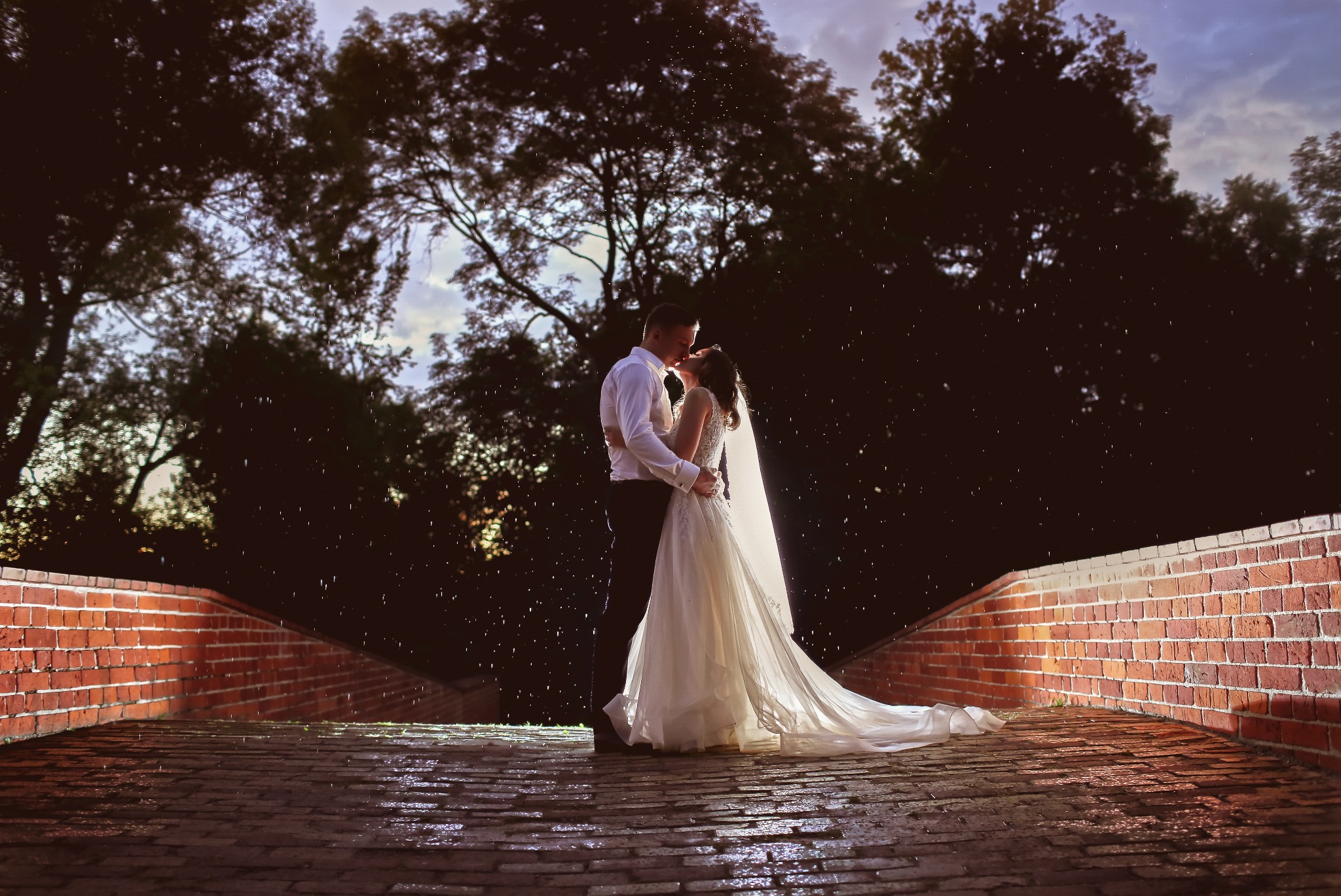 Para młoda w Ciechanowie całuje się na ceglanym moście w deszczu, drzewa w tle.