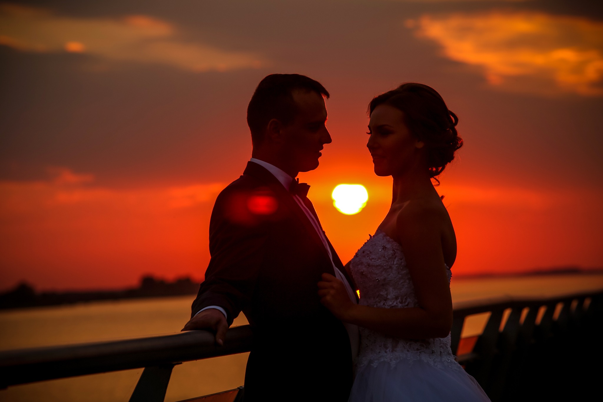 Para młoda w kontraście z zachodzącym słońcem nad wodą, ona w białej sukni ślubnej z koronką, on w czarnym smokingu z muszką, opierają się o barierkę.