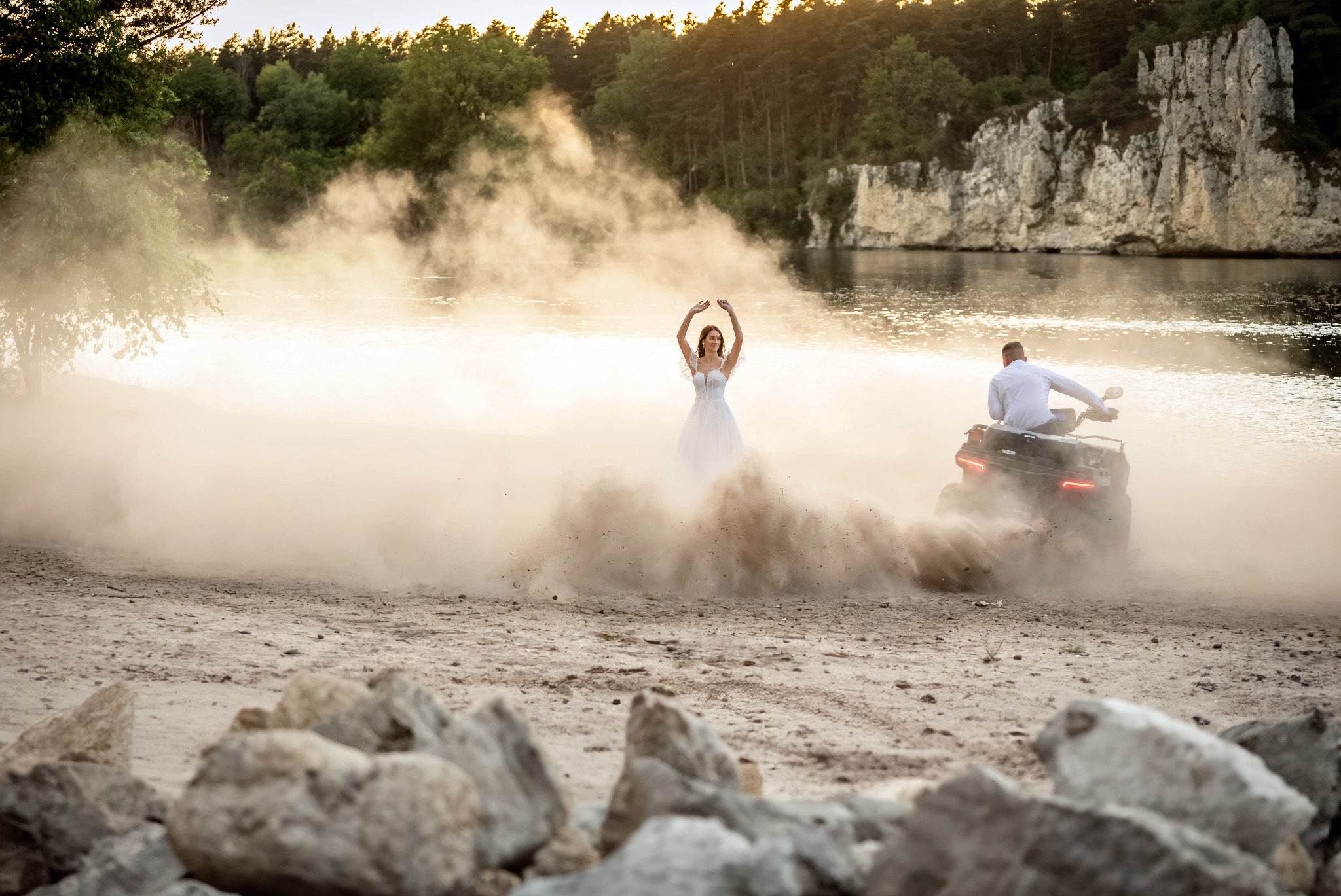 Panna młoda w białej sukni pozuje na piaszczystej plaży, otoczona tumami kurzu wznieconymi przez quada, którym jedzie pan młody w białej koszuli; w tle woda i skaliste klify.