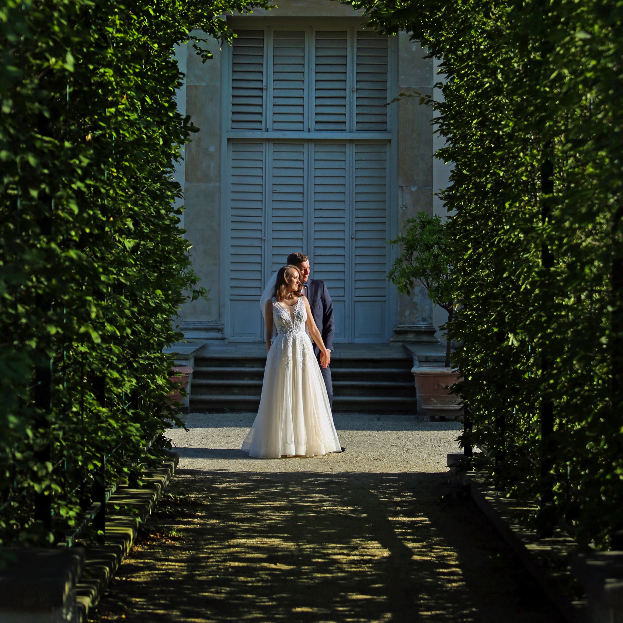 Para młoda w ogrodzie, panna młoda w białej sukni z koronkową górą, pan młody w granatowym garniturze, sfotografowani w alei z zielonych żywopłotów prowadzącej do budynku z szarymi okiennicami.