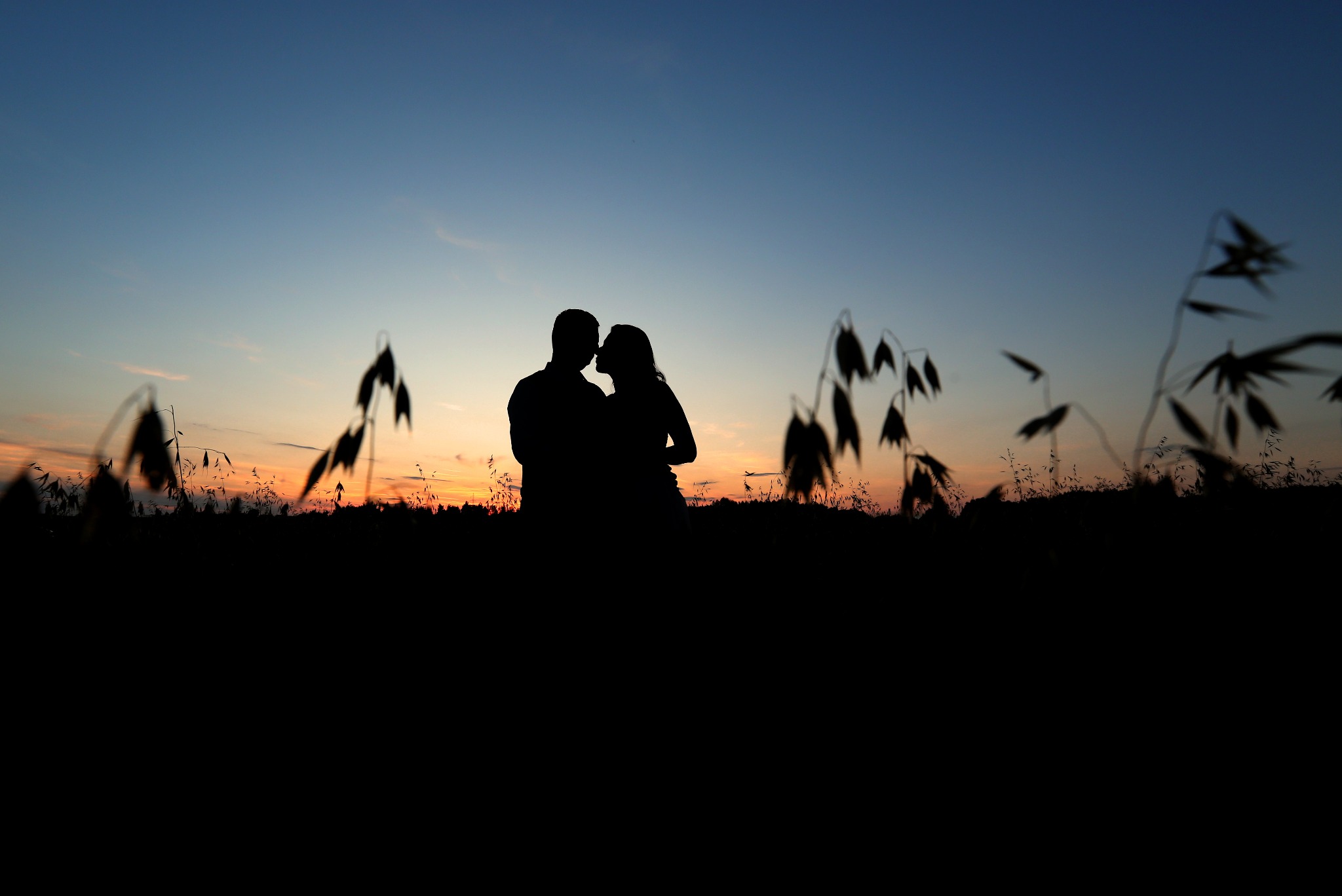 Sylwetka pary całującej się na tle zachodzącego słońca, zarysowana na tle nieba w odcieniach pomarańczy i błękitu, z delikatnymi źdźbłami trawy na pierwszym planie.