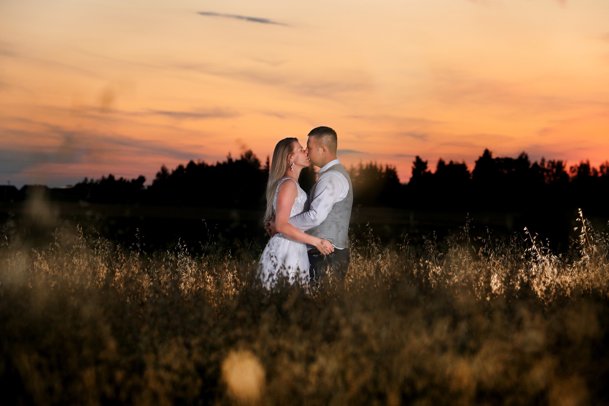 Para całująca się na tle zachodzącego słońca, otoczona wysoką trawą.