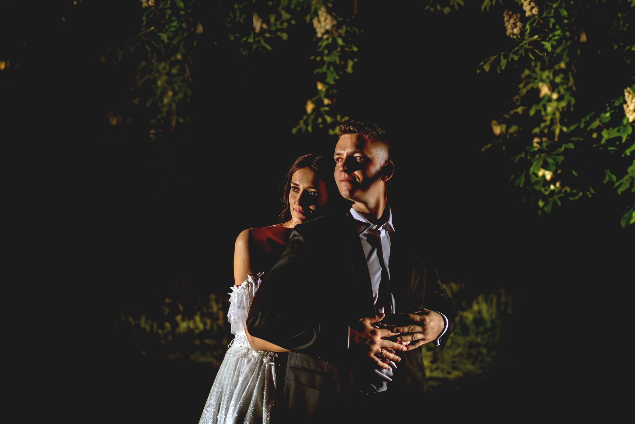 Para młoda w objęciach, on w czarnym garniturze, ona w błyszczącej białej sukni z koronkowymi ramionami, oświetleni punktowym światłem na tle ciemnej zieleni.