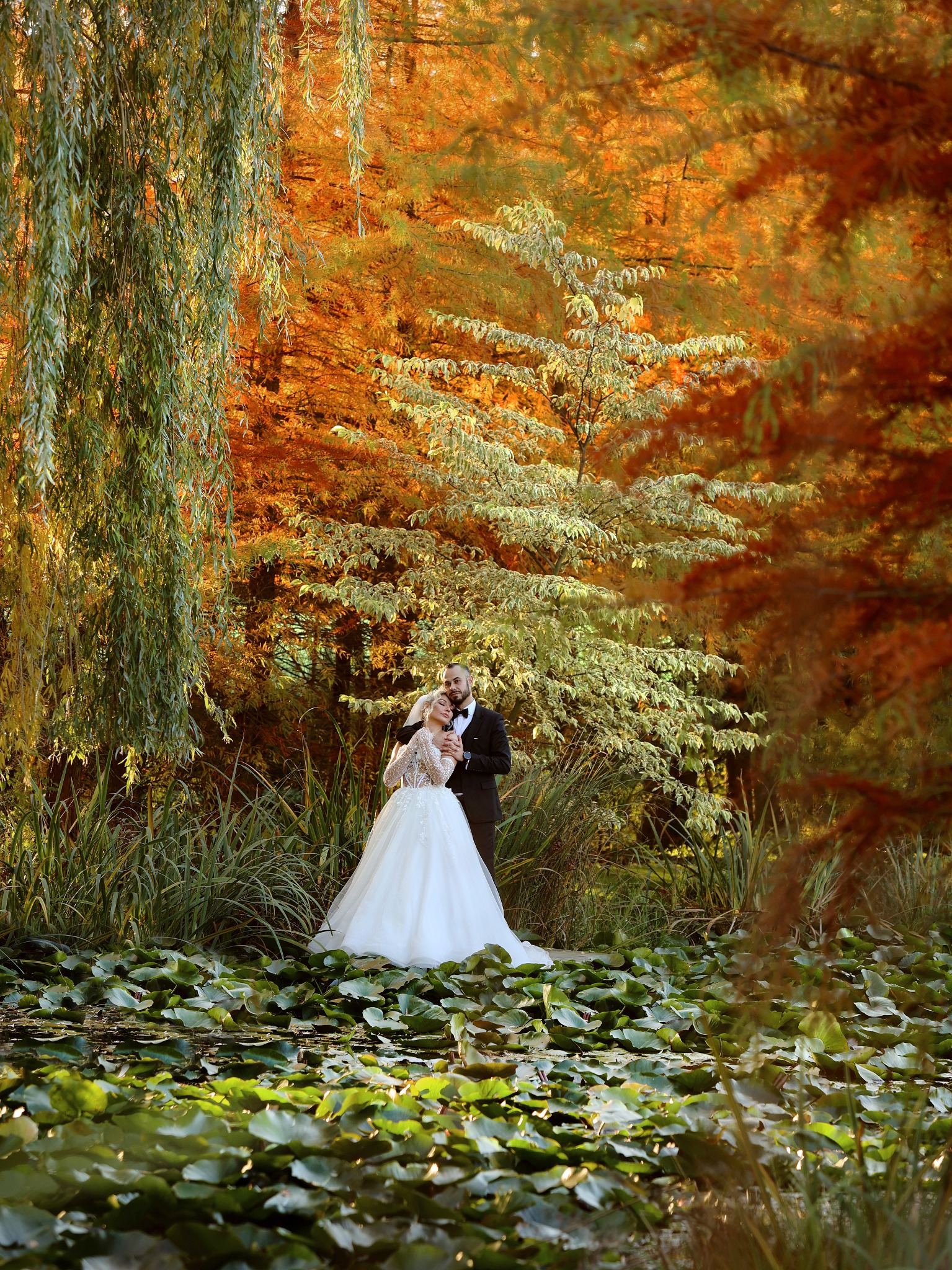 Para młoda w jesiennej scenerii nad stawem z liliami wodnymi, panna młoda w białej sukni z koronkowymi rękawami, pan młody w czarnym smokingu, drzewa w tle w odcieniach zieleni i pomarańczy.