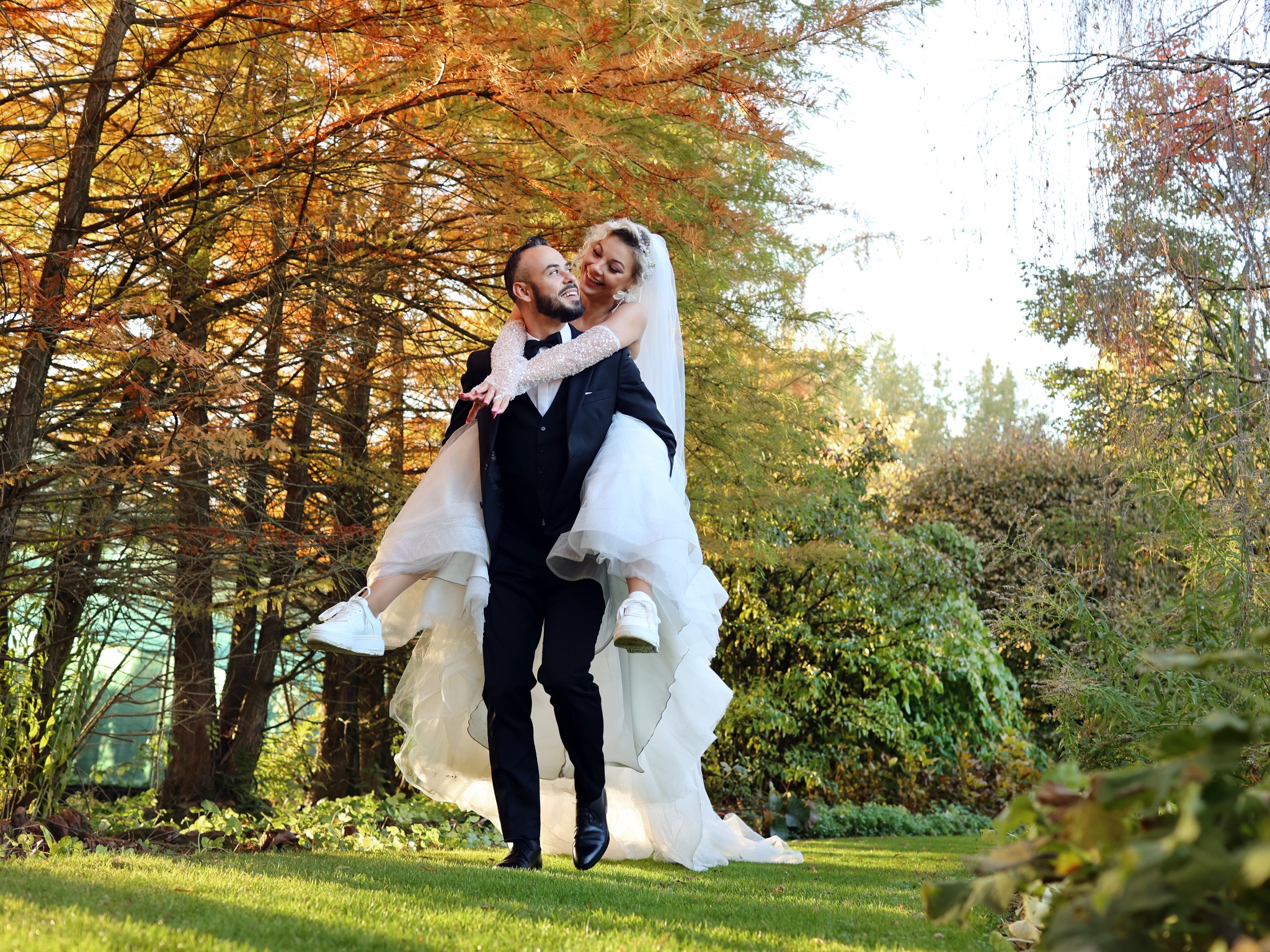 Uśmiechnięta panna młoda w białej sukni i sportowych butach na barana u pana młodego w czarnym smokingu, jesienne drzewa w tle.