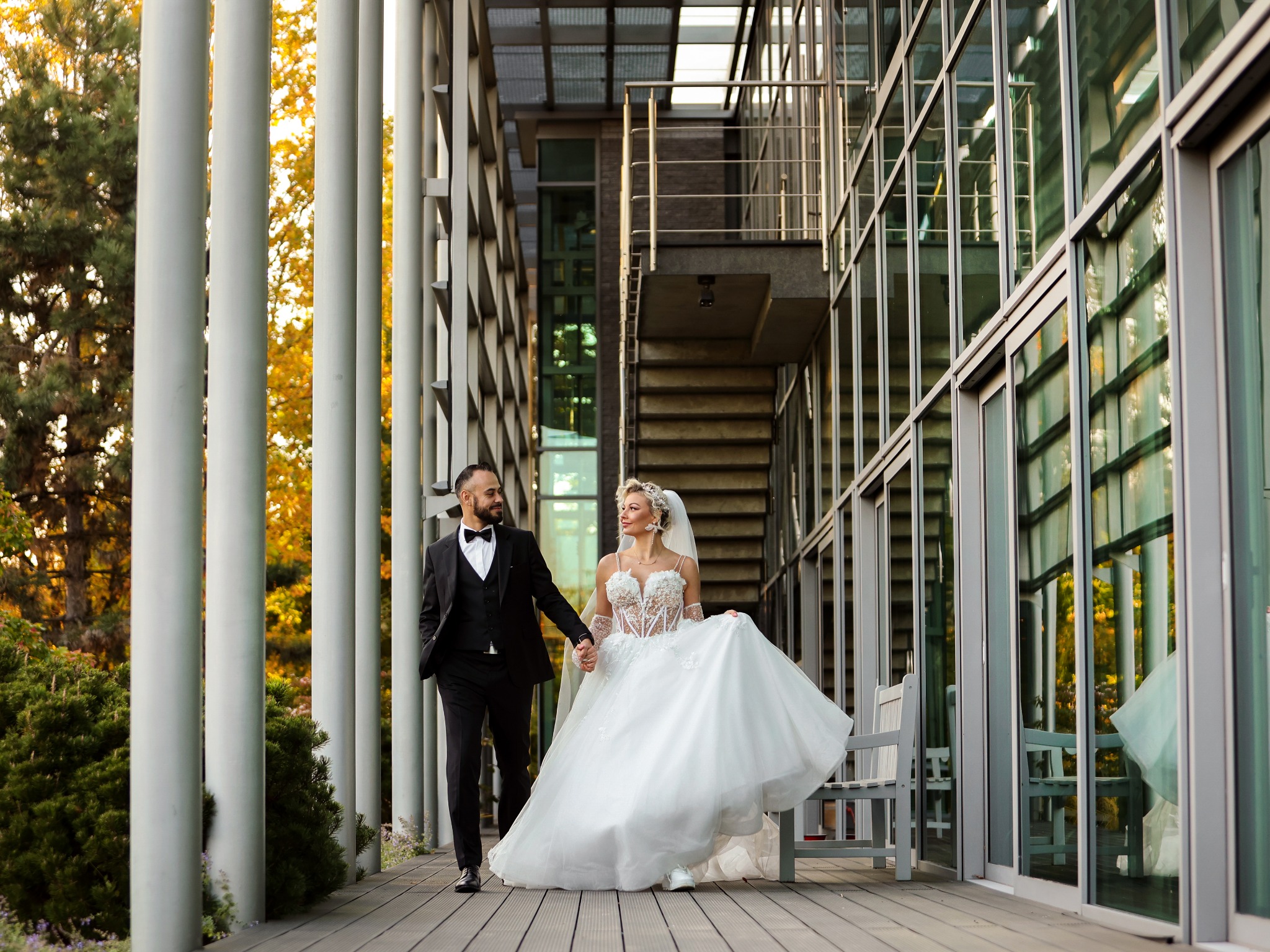 Para młoda, mężczyzna w czarnym garniturze i kobieta w białej sukni ślubnej z koronkowym gorsetem, spacerują trzymając się za ręce na tle nowoczesnej architektury z metalowymi kolumnami...