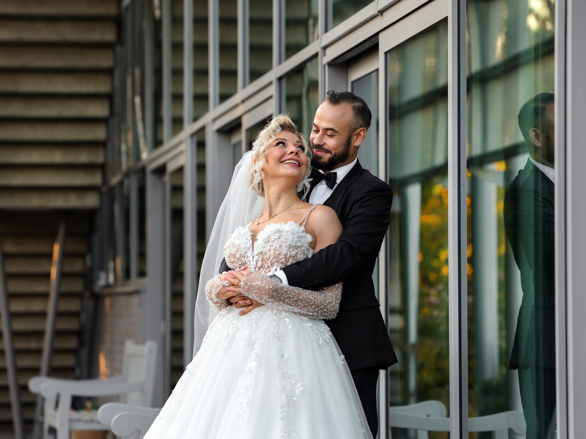 Uśmiechnięta para młoda w objęciach na tle nowoczesnej architektury z przeszkloną fasadą; panna młoda w bogato zdobionej sukni z koronkową górą i welonem, pan młody w czarnym smokingu z muszką.