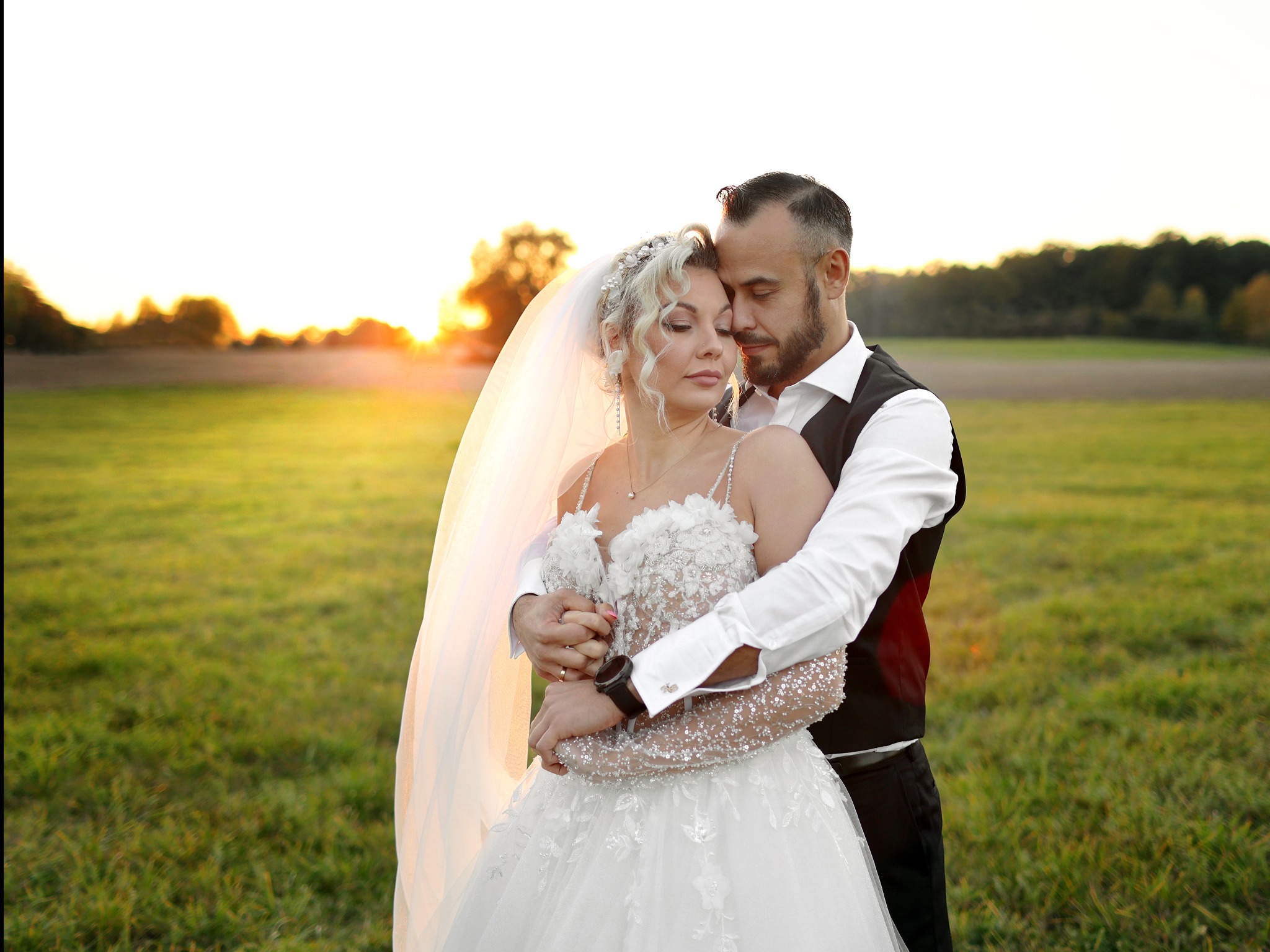 Para młoda przytulona na łące podczas zachodu słońca, panna młoda w bogato zdobionej sukni z welonem, pan młody w czarnej kamizelce, delikatne oświetlenie podkreśla romantyczny nastrój.