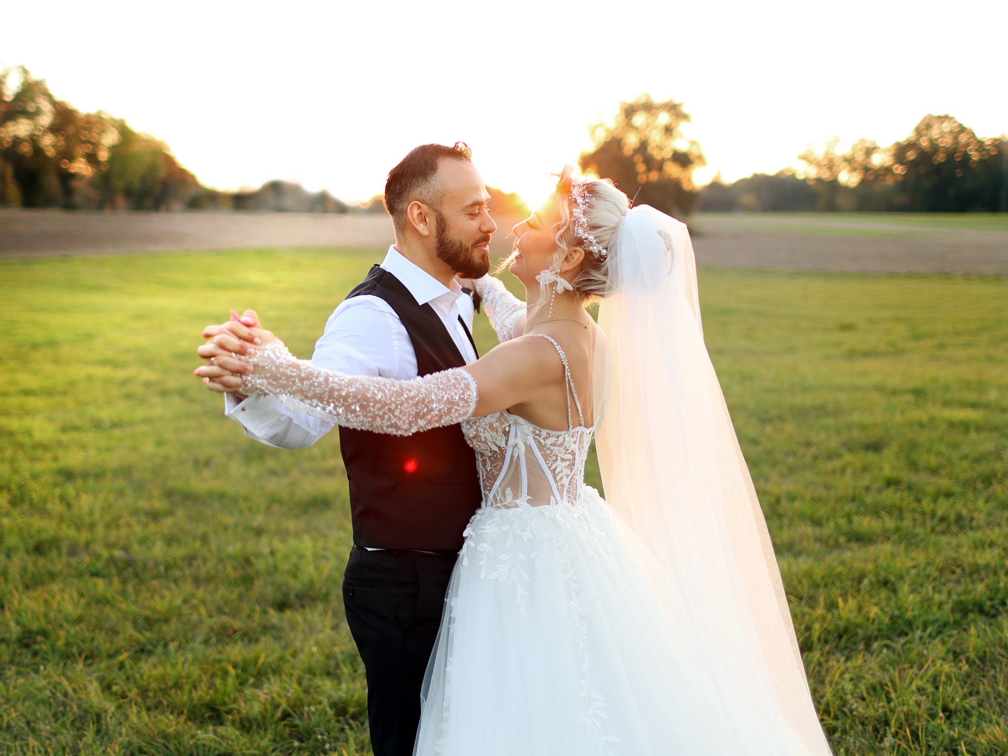 Para młoda w tańcu na tle zachodzącego słońca w plenerze, ona w białej sukni z koronkowymi rękawami, on w czarnej kamizelce i białej koszuli.