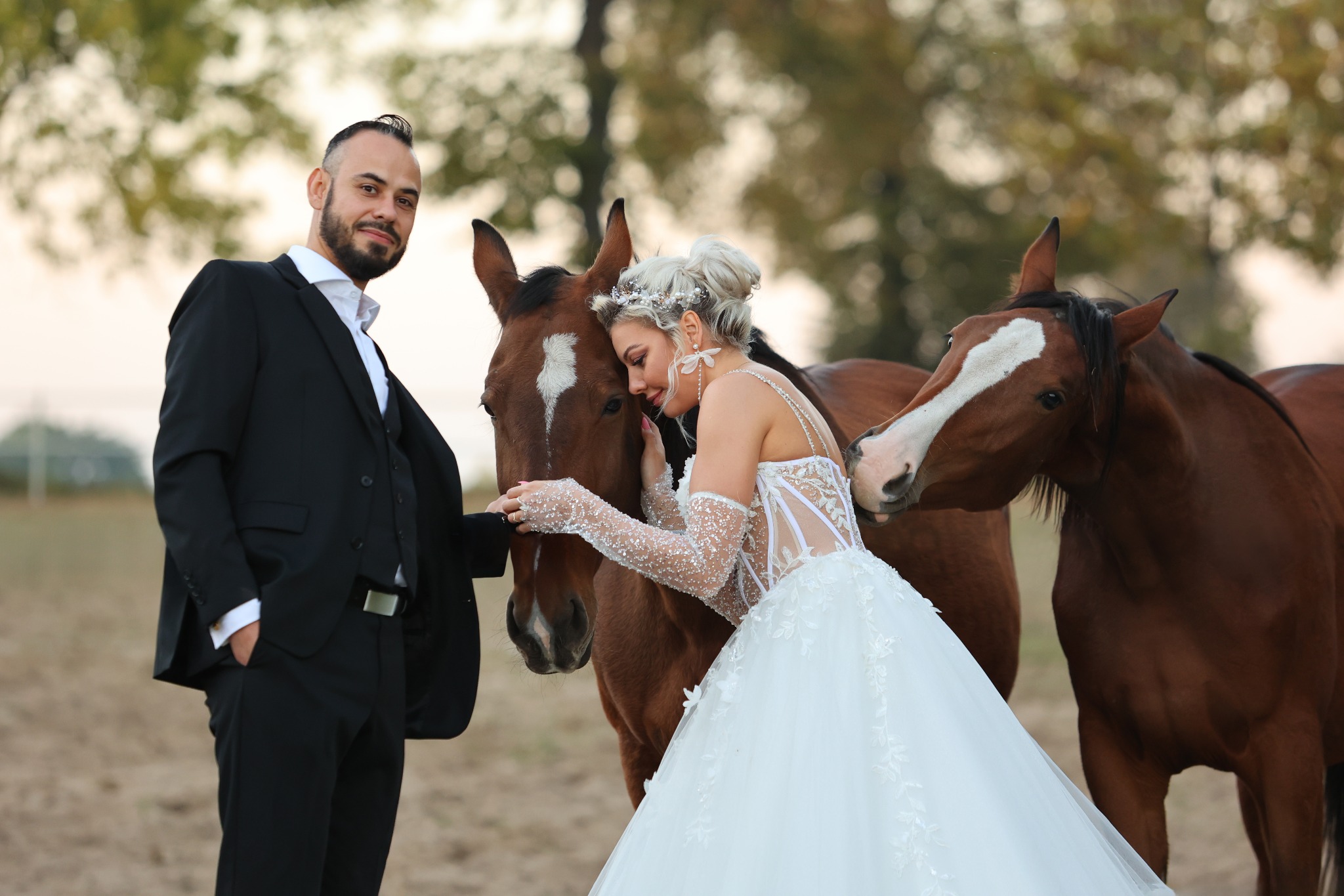Elegancko ubrana para pozuje z dwoma brązowymi końmi na tle drzew i pola; panna młoda w sukni z koronkowymi rękawami opiera głowę o konia.