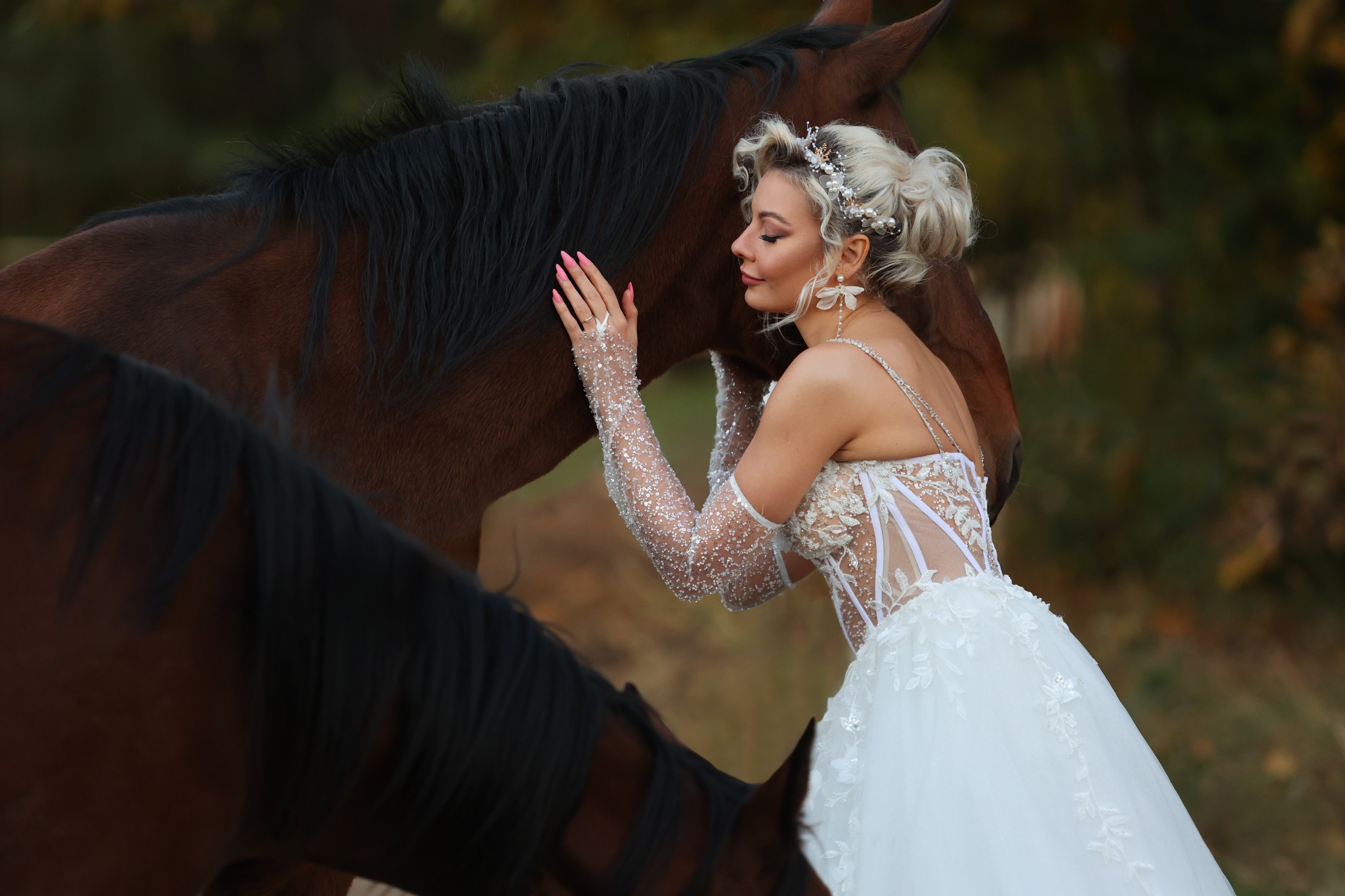 Panna młoda w białej sukni ślubnej z koronkowymi rękawami, ozdobną fryzurą i kwiatowymi kolczykami, głaszcze brązowego konia, z drugim koniem w tle.