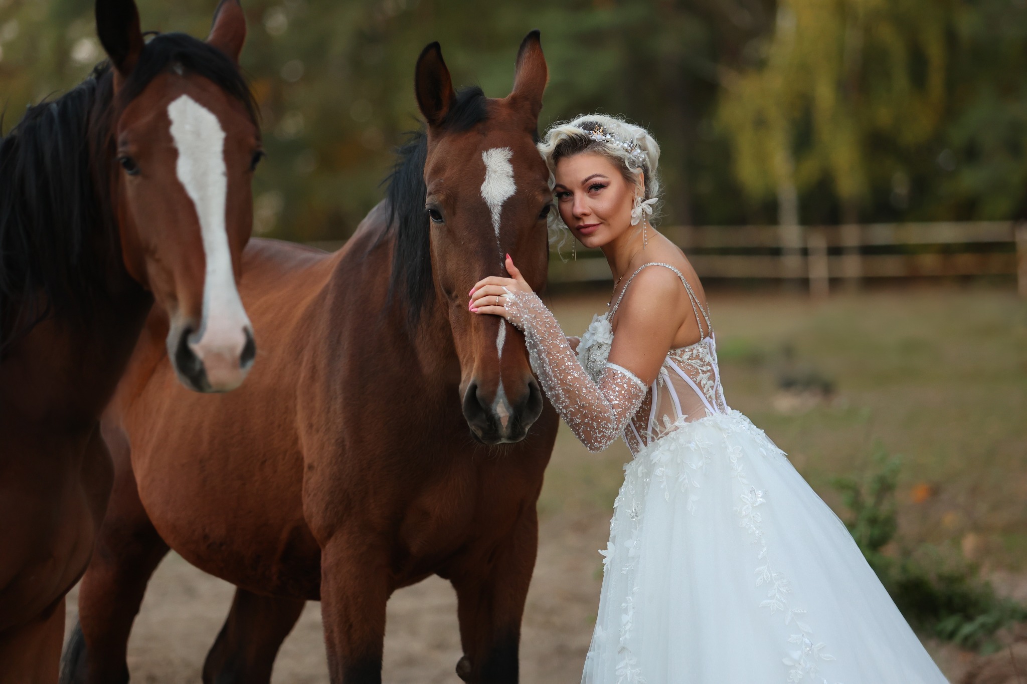 Panna młoda w białej sukni z koronkowymi rękawami, pozująca z dwoma brązowymi końmi z białymi strzałkami na głowach, w plenerze.