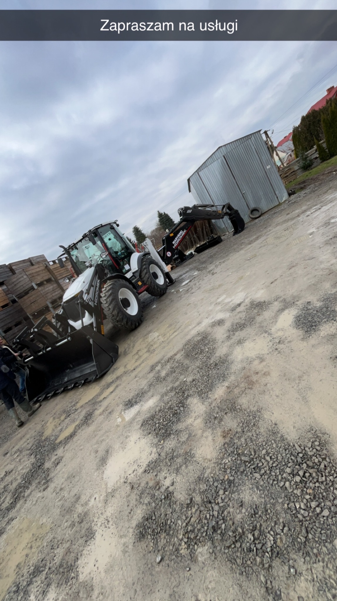 Biało-czarna koparko-ładowarka marki Hidromek na grząskim, szutrowym terenie, z widocznym stosem drewnianych skrzyń i blaszakiem w tle, pochmurne niebo.