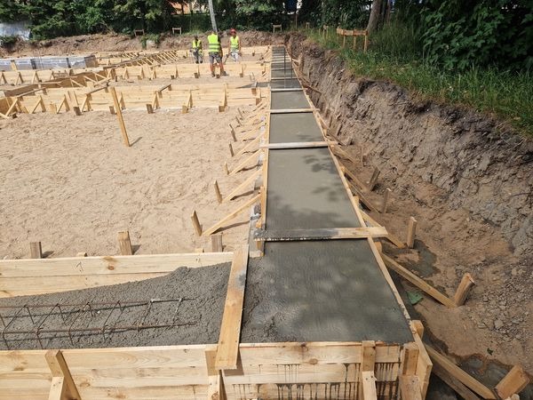Wykonywanie fundamentów: zalewanie betonem szalunków zbrojeniowych, widoczni pracownicy w odblaskowych kamizelkach w tle.