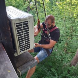 GooDoor - Mężczyzna w uprzęży alpinistycznej montuje zewnętrzną jednostkę klimatyzacji na wysokości, z widocznym otoczeniem zieleni.