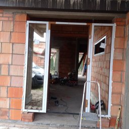 GooDoor - Montaż okna w surowym budynku z czerwonej cegły, widoczna drabina, wiertarka udarowa i poziomica, laserowy wskaźnik poziomu, na zewnątrz zaparkowany samochód dostawczy.