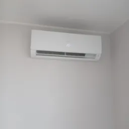 Biały klimatyzator Haier zamontowany na jasnoszarej ścianie w rogu pokoju, wyświetlający temperaturę 20 stopni Celsjusza.