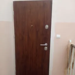 Ciemnobrązowe drzwi wejściowe z srebrną klamką i wizjerem, zamontowane w bloku mieszkalnym, widoczne ślady po montażu na ścianie, w tle fragment grzejnika i puszka farby.