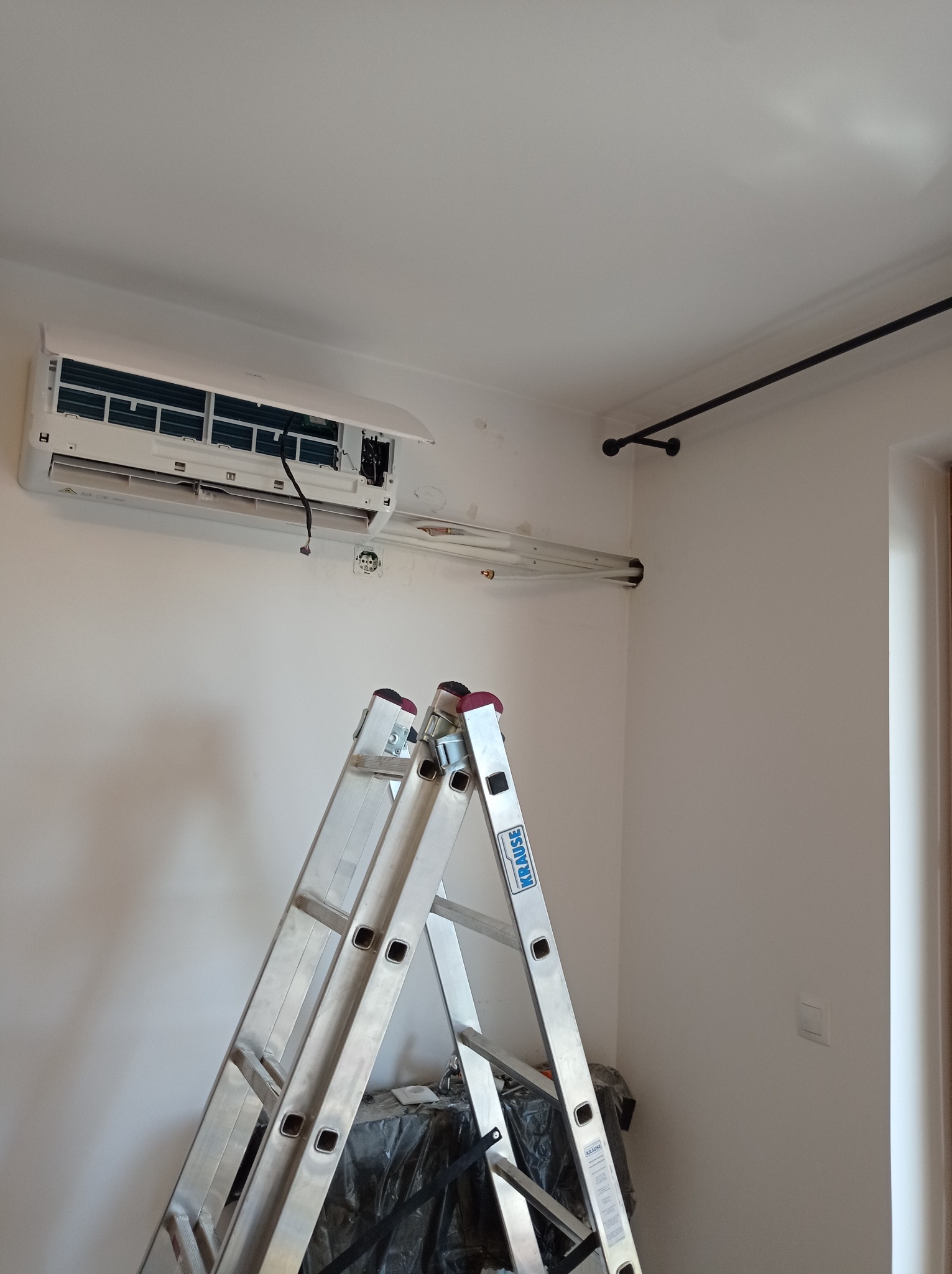 Biały klimatyzator ścienny z otwartą klapą i widocznymi elementami wewnętrznymi, zamontowany na białej ścianie w trakcie instalacji, pod nim aluminiowa drabina Krause i zabezpieczona folią podłoga.