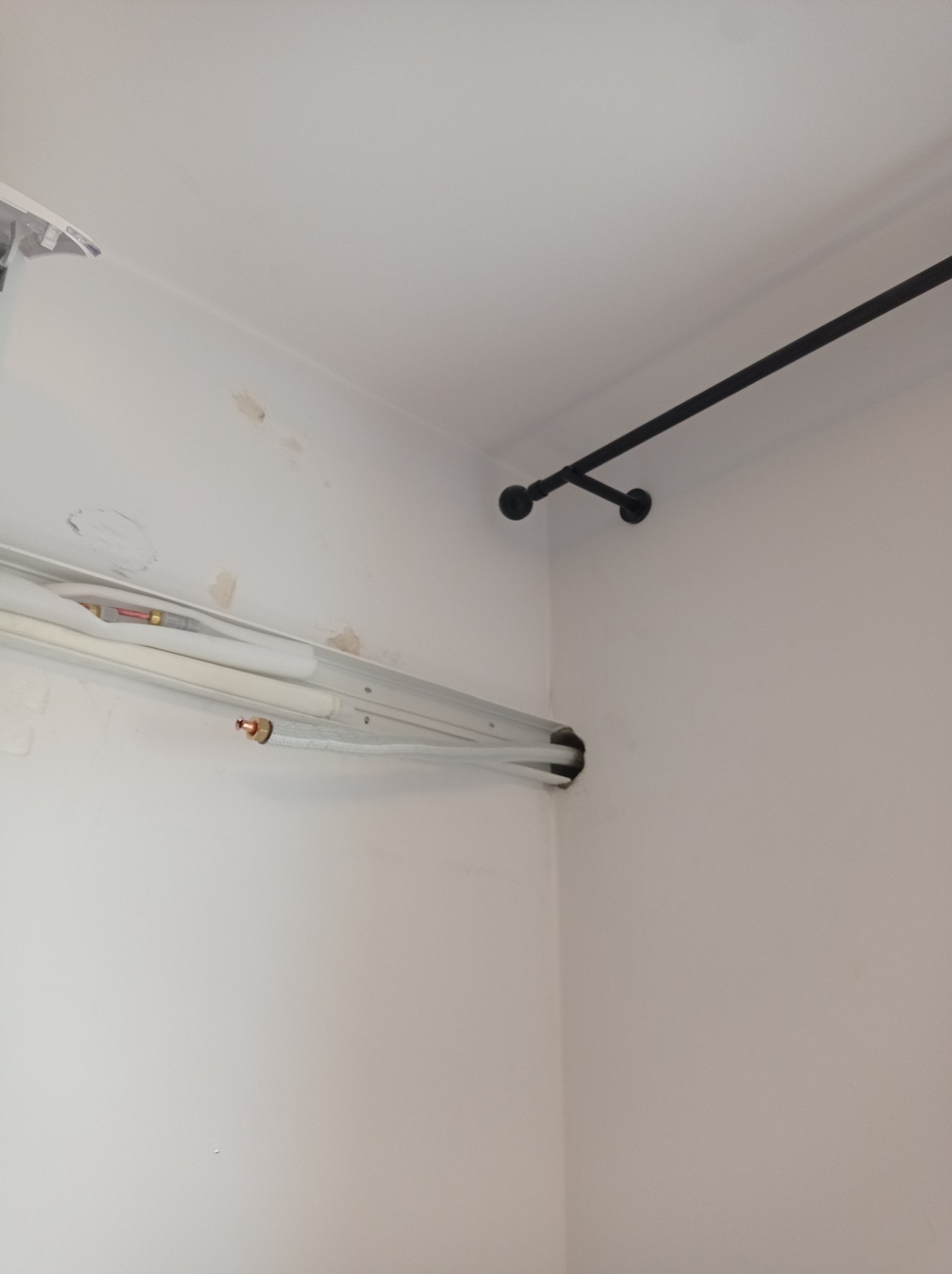 Rury instalacyjne, prawdopodobnie klimatyzacji, prowadzone wzdłuż ściany pod sufitem, z widocznym otworem w rogu pomieszczenia i czarnym, metalowym drążkiem zamontowanym powyżej.